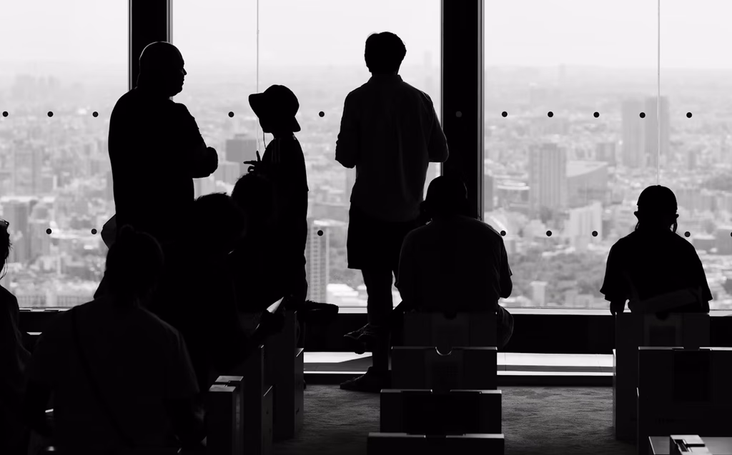 Silhouettes of people standing and sitting near a large window overlooking a cityscape. The scene is in black and white, with the figures in shadow and the city skyline visible through the glass.